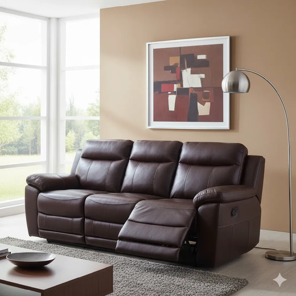 A comfortable three-seater brown leather reclining sofa with plush cushions and visible reclining mechanisms on the far right and left seats, where the footrests are partially extended. The sofa is situated on a light gray shag rug in a bright living room. The walls are painted a warm beige, and a large abstract art piece with earthy tones of brown, red, and white hangs framed above the sofa. A large window on the left offers a view of green trees outside. To the right, a silver arc floor lamp with a dome-shaped shade provides lighting. A modern wooden coffee table with a dark brown top and black legs is partially visible in the foreground, holding a decorative bowl and books.