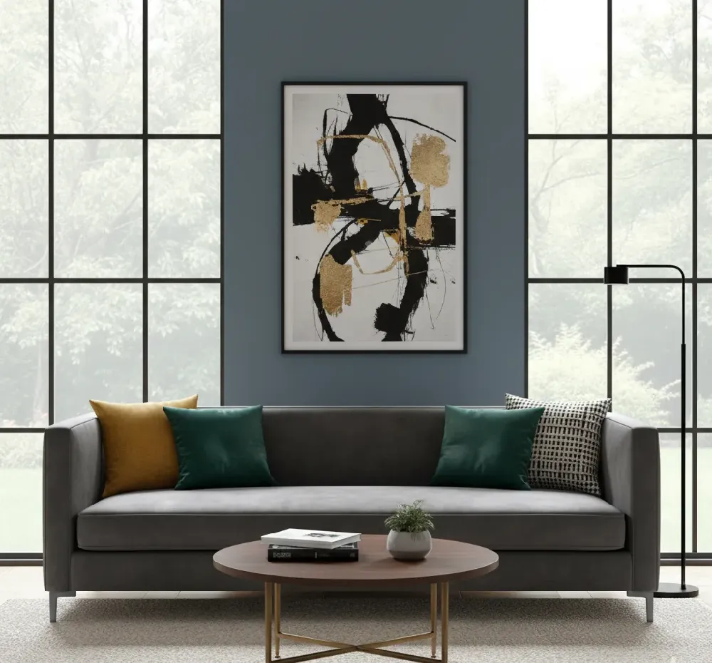 A sleek, modern charcoal gray Tuxedo sofa with straight arms and a back that are the same height, resting on thin metal legs. The sofa is adorned with throw pillows in mustard yellow, emerald green, and a black and white geometric pattern. In front of the sofa is a round wooden coffee table with a gold metal base, holding a small potted plant and some books. The sofa is placed on a light textured rug. The wall behind the sofa is painted a deep blue-gray, featuring a large framed abstract art piece with black, white, and gold brushstrokes. Large black-framed windows offer a view of green trees outside. A minimalist black floor lamp stands to the right of the sofa.